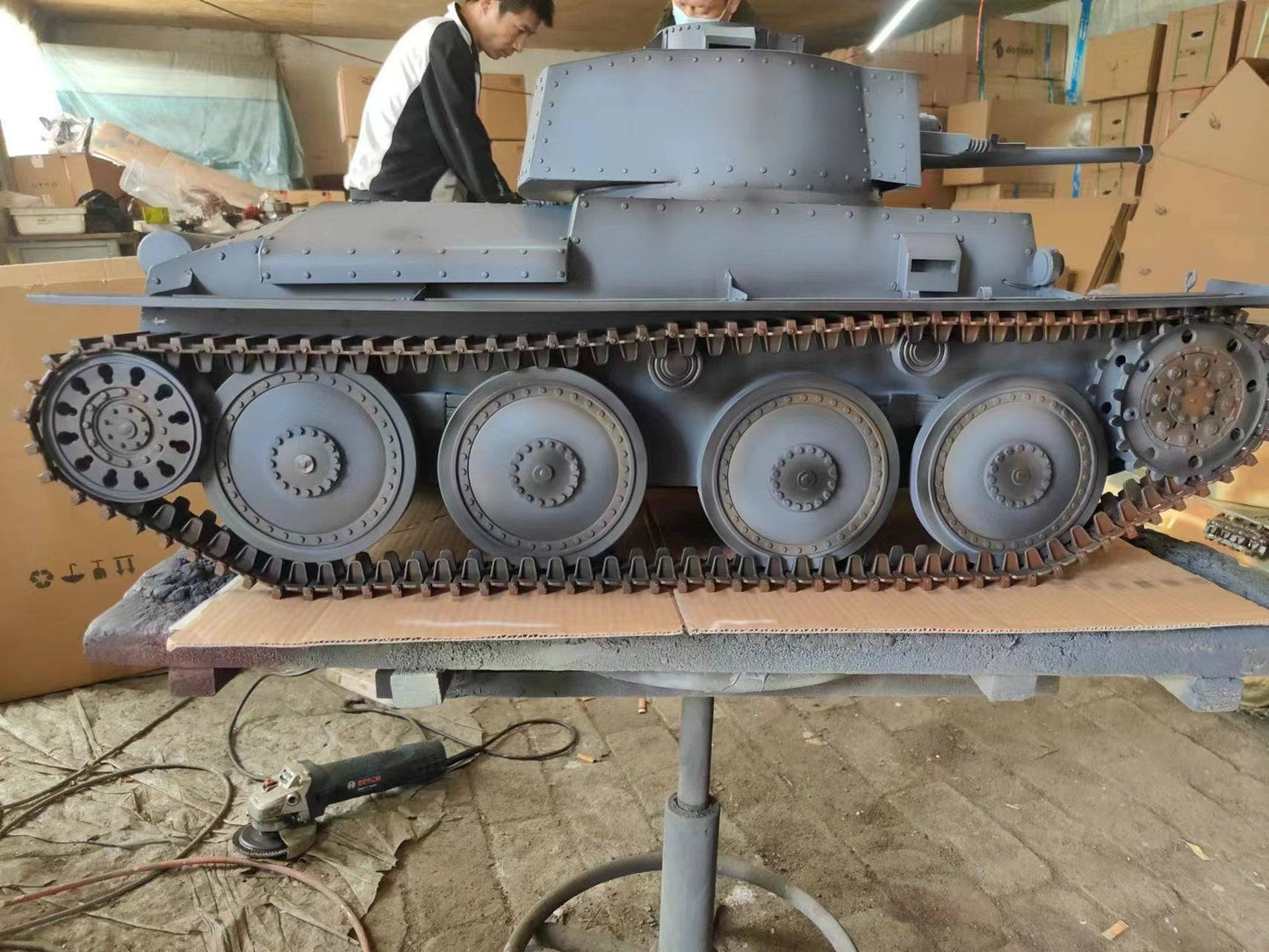 An eaxtoys 1/6 scale gray Panzerkampfwagen 38(t) model, mimicking a WWII German tank, is on display in a possible workshop or garage. Two military model enthusiasts are partially visible in the background, deeply focused on examining the detailed replica.
