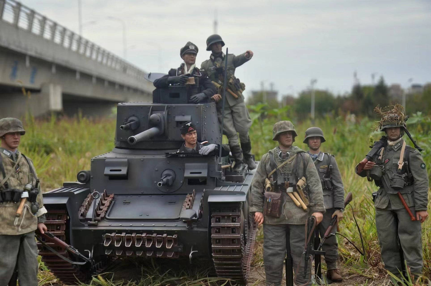 Six soldiers in WWII uniforms gather around the eaxtoys 1/6 scale Panzerkampfwagen 38(t) model tank in a grassy field. One soldier stands in the hatch while others position themselves around this iconic German tank. A bridge looms under the cloudy sky behind them.