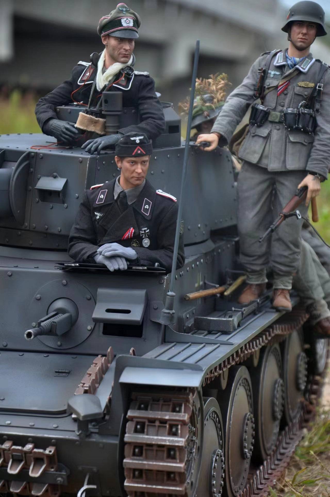 Three figures in historical uniforms are displayed on an eaxtoys 1/6 scale Panzerkampfwagen 38(t), a beloved WWII German tank model. The setup includes two standing figures, one with a rifle, and one seated in the tank, all set against a backdrop of greenery and a bridge.