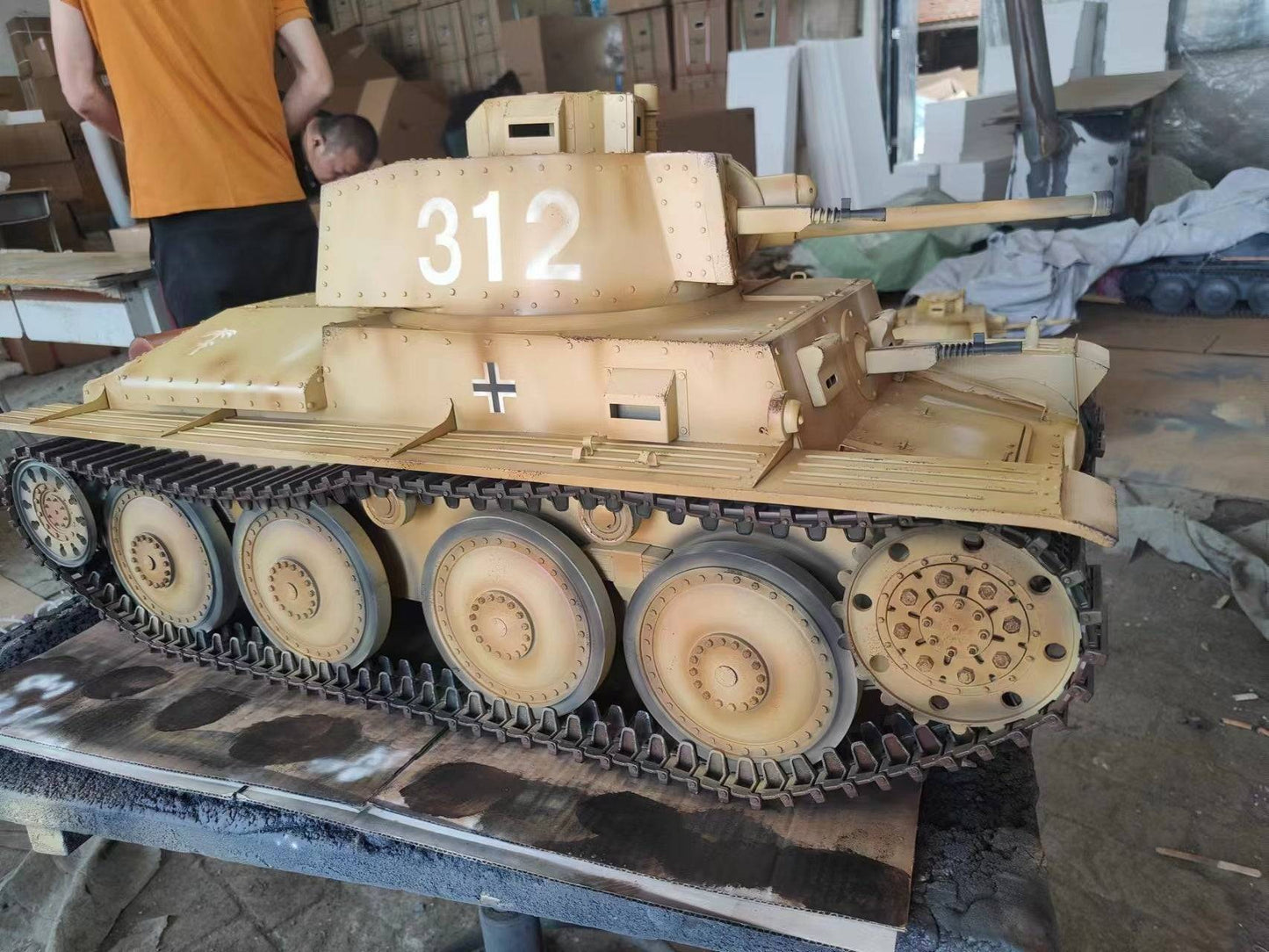 An eaxtoys 1/6 scale Panzerkampfwagen 38(t) model, detailed with 312 and a German cross, sits on a table. In the background, a bustling workshop teems with military model enthusiasts examining boxes while someone in an orange shirt diligently works.
